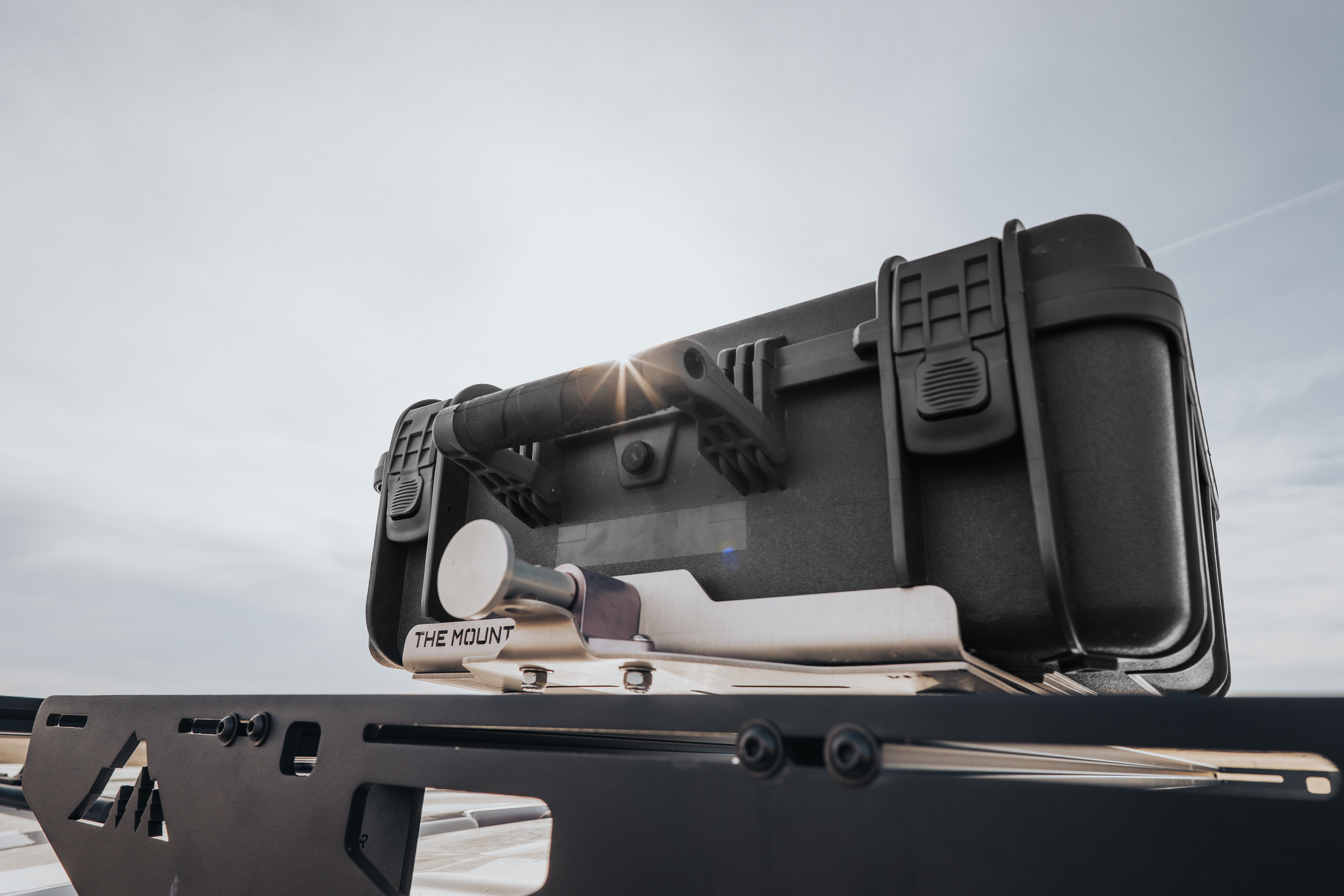 Black rugged protective case with secure latches and handle, displayed on a gun rack under cloudy sky.