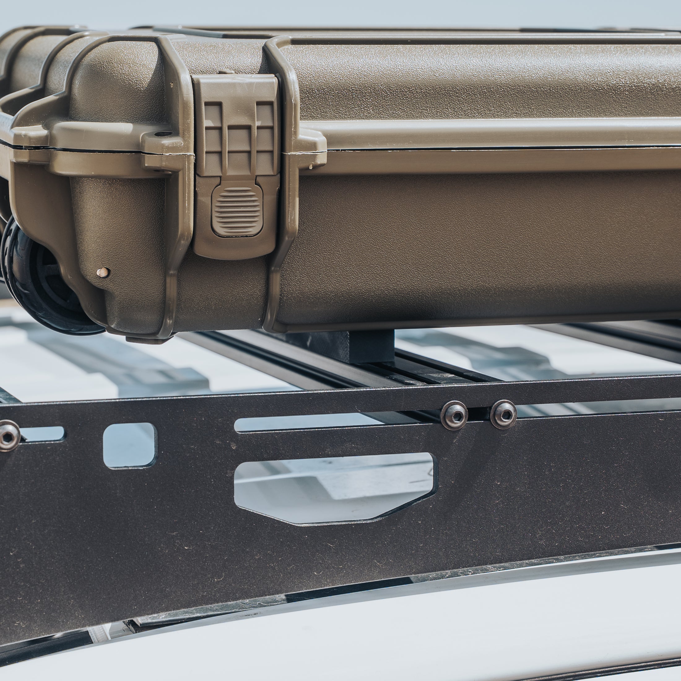Black metal case stabilizer mounted on vehicle roof rack supporting a tan hard plastic case with secure latches.