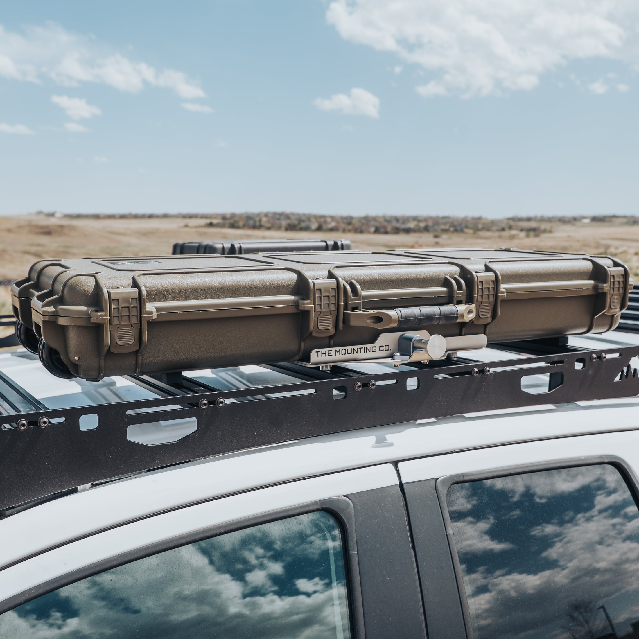 Durable olive green hard case with reinforced latches mounted on black roof rack of white vehicle.