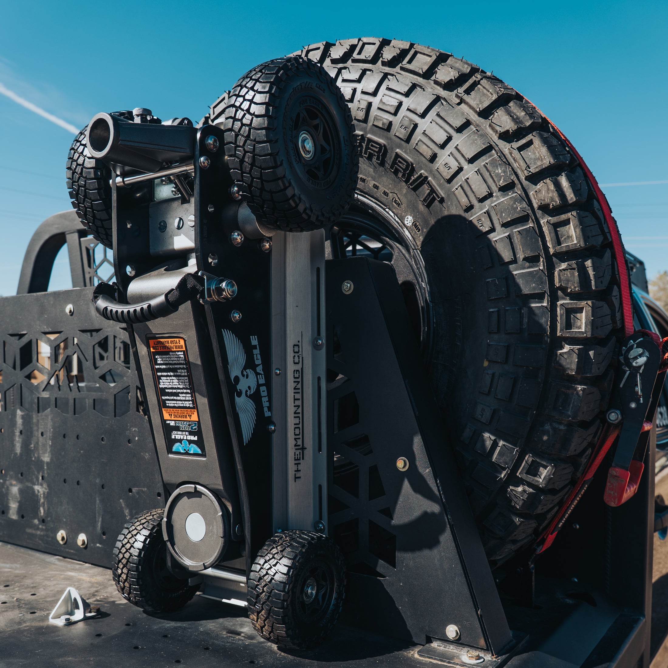 Black steel Pro Eagle 2 Ton jack mounted next to large spare off-road tire on vehicle rack.
