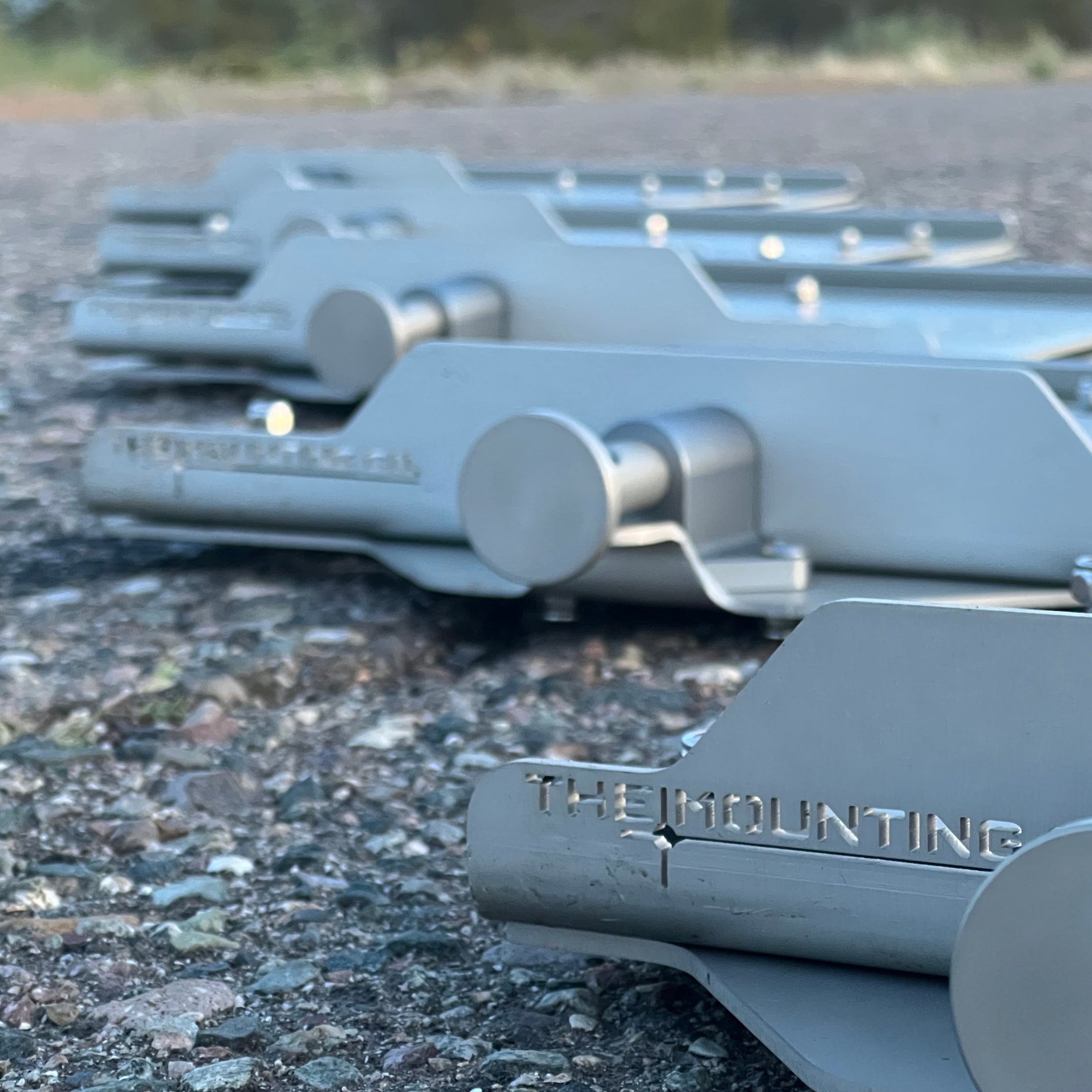 Stack of matte silver aluminum mounts with rounded ends and engraved "THE MOUNTING" logo on rough ground surface.