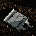 Load image into Gallery viewer, Compact silver aluminum mount plate with black screws and a quick-release lever on dark soil background.
