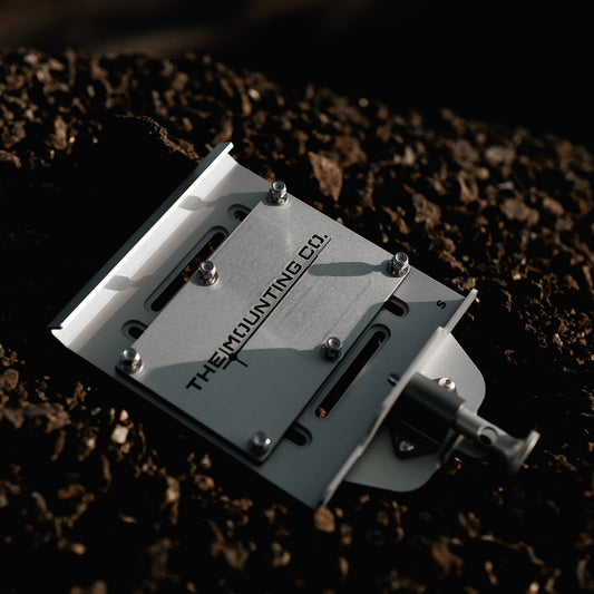 Compact silver aluminum mount plate with black screws and a quick-release lever on dark soil background.